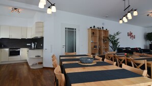 Cocina moderna con armarios blancos, encimera negra y horno empotrado.  Hay un comedor con mesa y sillas de madera, además de una estantería llena de libros y objetos decorativos.