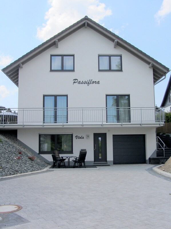 A two-story building named Passiflora with a balcony, a garage, and a seating area with chairs.