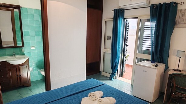 Une chambre avec un lit, un petit bureau, un réfrigérateur, une salle de bain avec un lavabo et un miroir, et une vue sur l’extérieur par une porte.