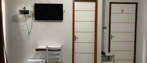 Una habitación con un televisor montado en la pared, un armario blanco, una silla blanca, un refrigerador blanco y dos puertas con un letrero de 'No Entrar'.
