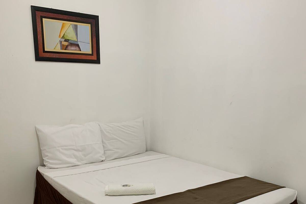 Una cama individual con base color café, ropa de cama blanca y una almohada blanca. Encima de la cama, un cuadro enmarcado cuelga de la pared.