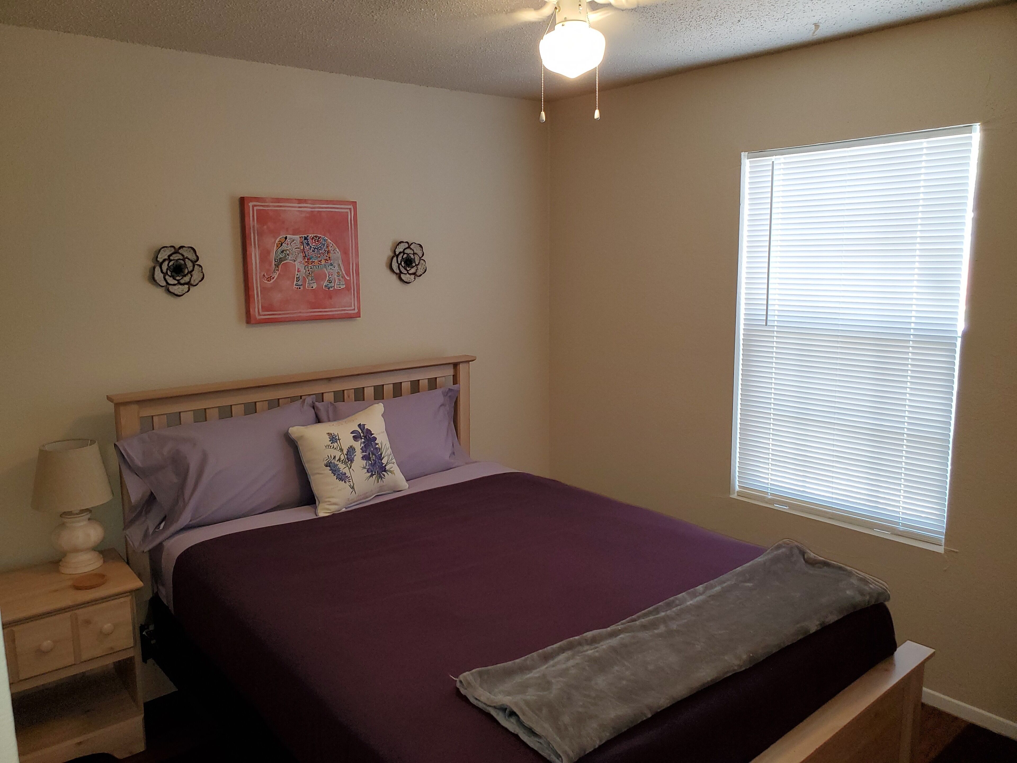 A bedroom with a bed, a nightstand, a lamp, a window with blinds, and two wall decorations.