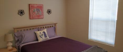 A bedroom with a bed, a nightstand, a lamp, a window with blinds, and two wall decorations.