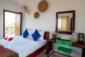 A hotel room with a bed, a desk, a chair, a fan, a mirror, and a window with curtains.