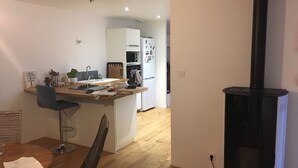 Ett modernt kök med en central köksö, en matplats med bord och stolar samt en svart vedkamin.