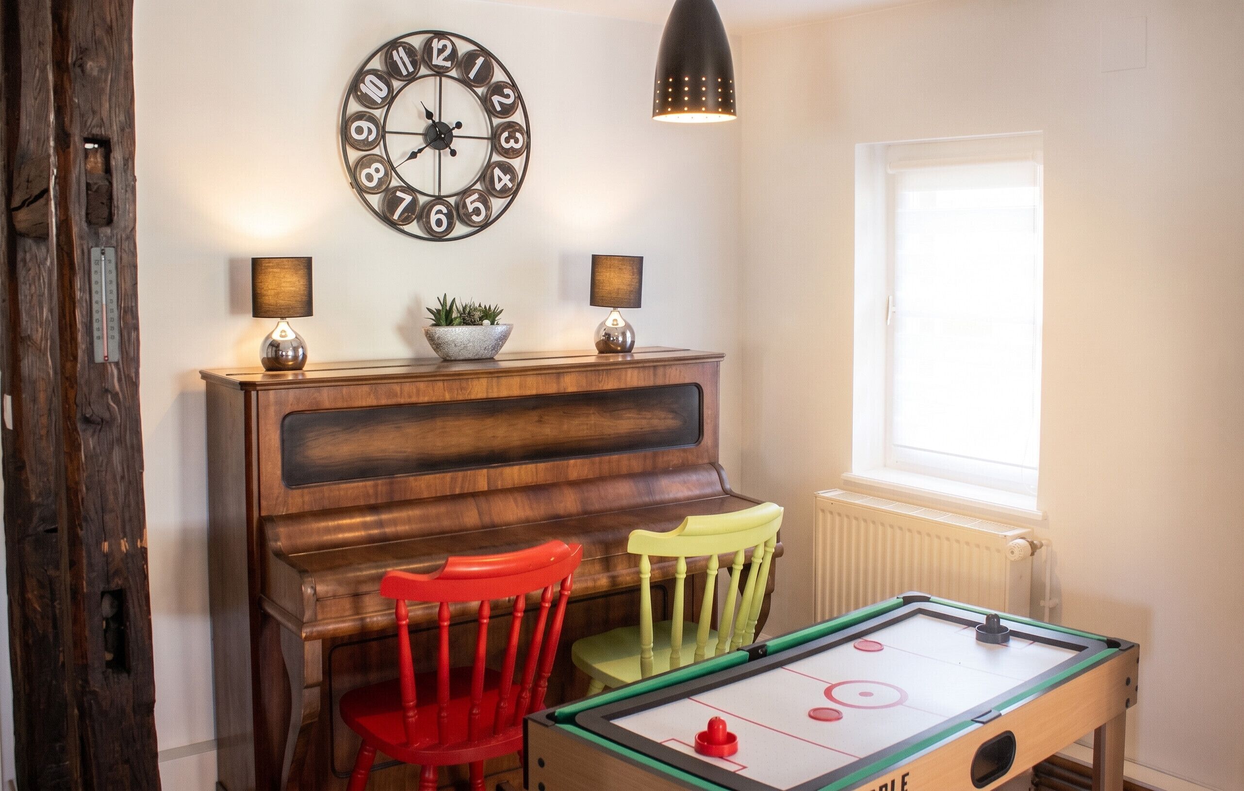 Una habitación con un piano de madera, dos sillas, una mesa de fútbol de mesa y un reloj de pared, además de una ventana.