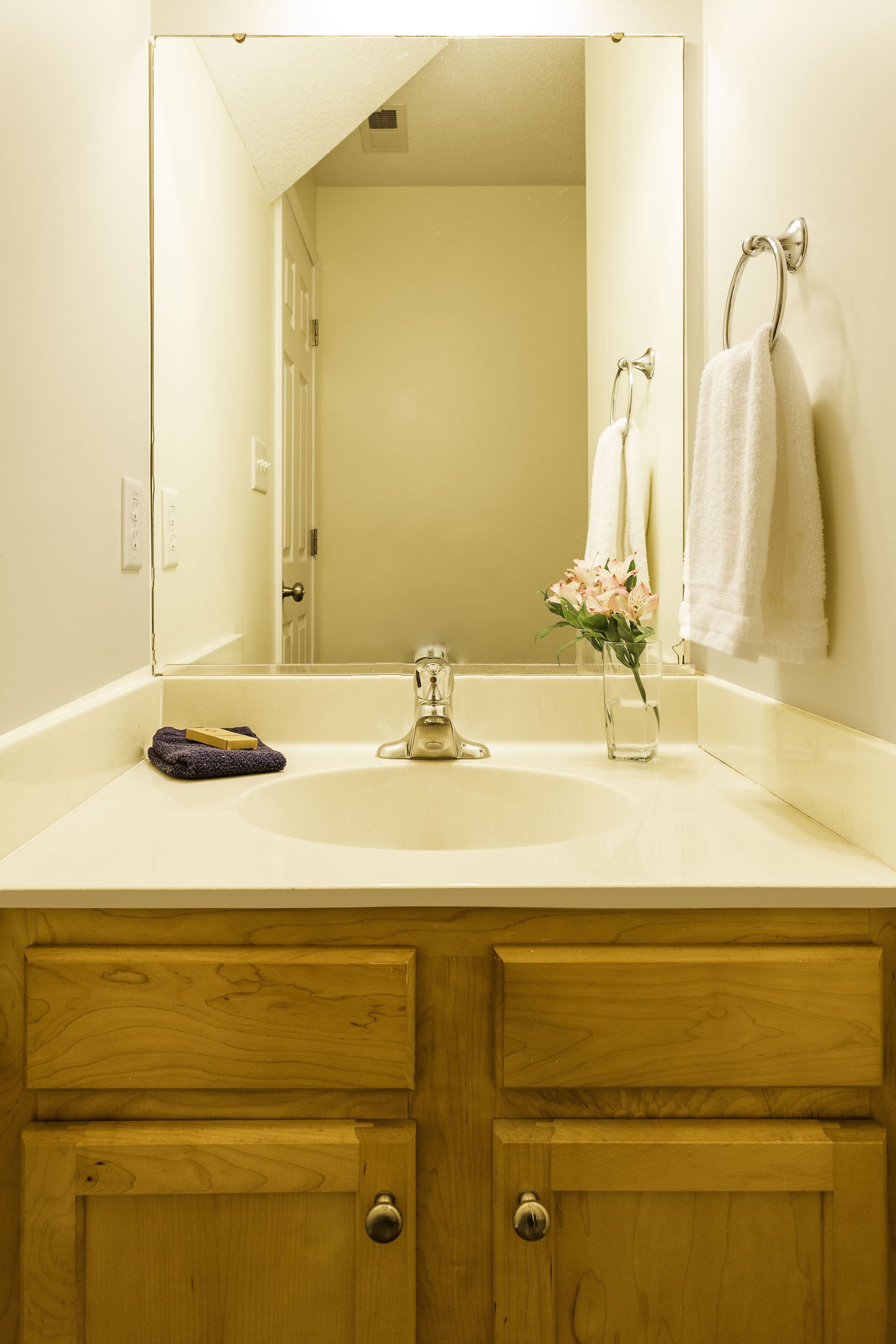 A bathroom with a white sink, wooden vanity, and a mirror reflecting a towel and a door.
