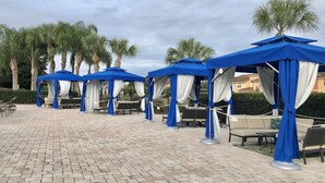 Varias zonas de estar al aire libre con toldos azules y cortinas blancas, dispuestas en un patio empedrado con palmeras al fondo.