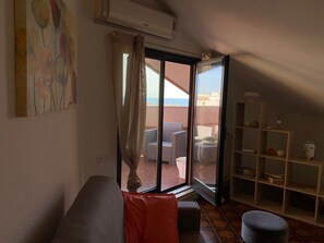 Ein Wohnzimmer mit einer Couch, einem roten Kissen, einer Glastür, die auf einen Balkon führt, und einem Bücherregal.