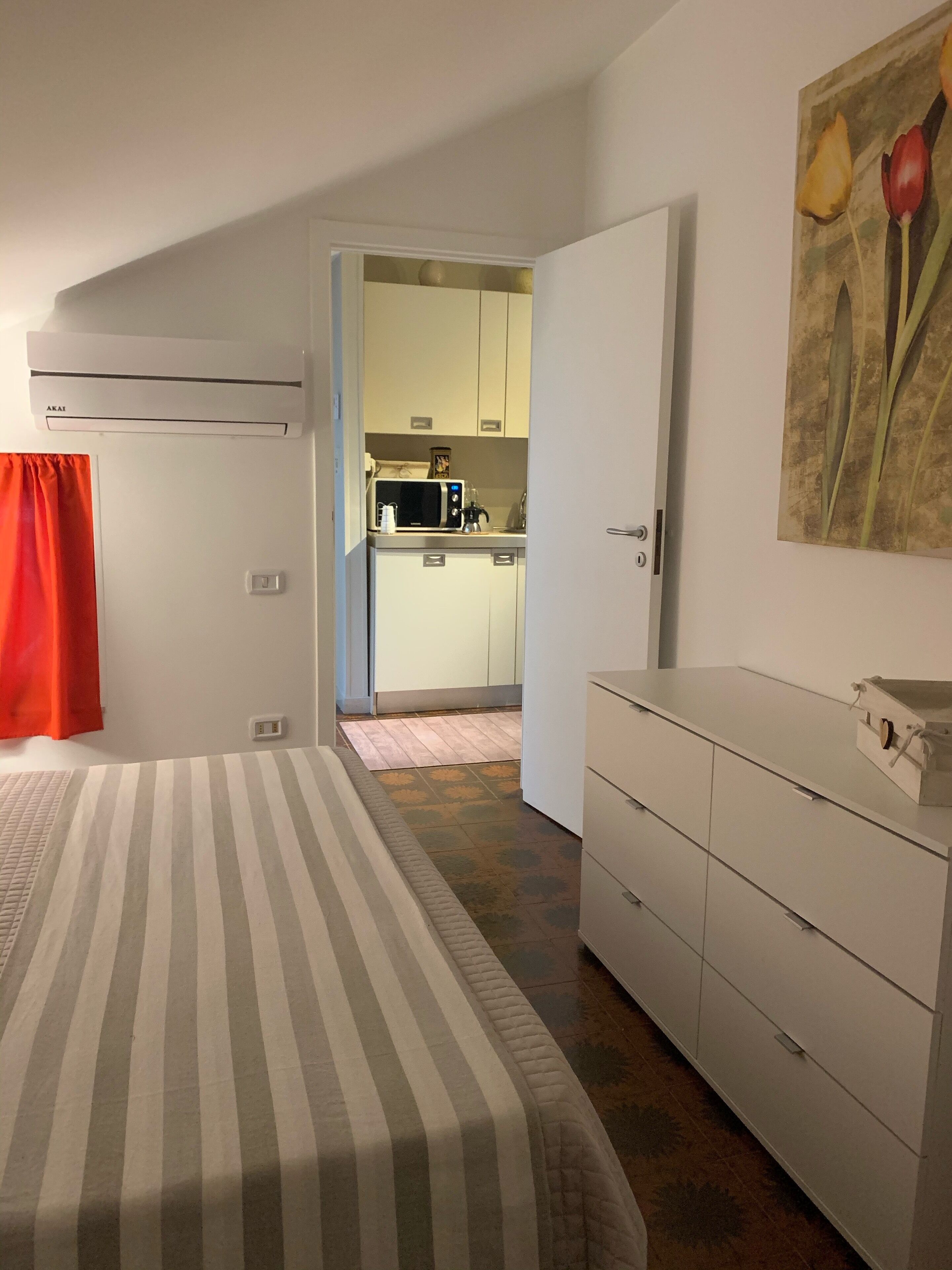 A bedroom with a bed, a dresser, and a view into the kitchen.