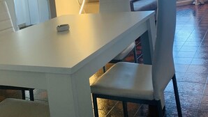 Un comedor con una mesa y sillas blancas, dos cuadros en la pared y vistas al exterior a través de una ventana con cortinas.