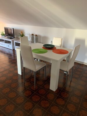 Ein Essbereich mit einem weißen Tisch, vier Stühlen, einem Fernseher auf einem Schrank und dekorativen Elementen an der Wand.