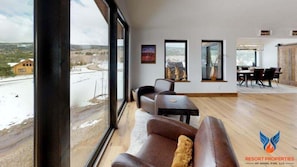 A spacious room with a large window offering a view of a river and distant buildings. There is a seating area with a sofa and a table, and a dining area with a table and chairs.
