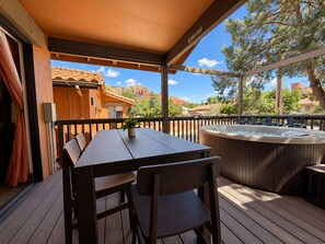 Une terrasse en bois avec un jacuzzi, une table et des chaises, offrant une vue panoramique sur un paysage verdoyant, avec des arbres et un bâtiment en arrière-plan.