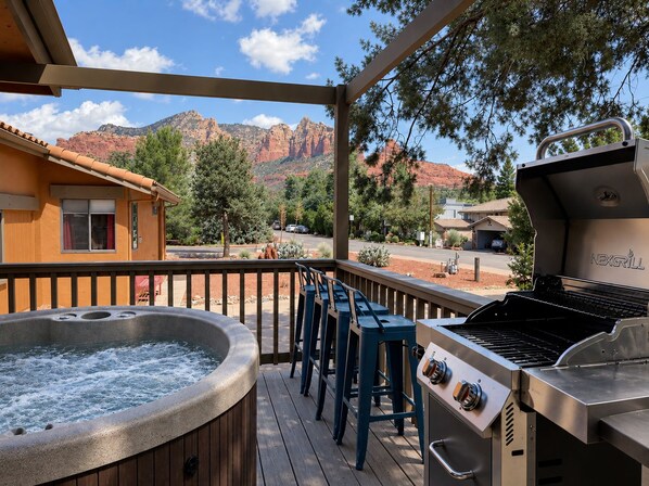 A hot tub, bar stools, and a grill on a wooden deck with a mountain view.