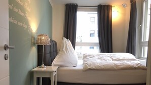 Un dormitorio con cama, una mesita de noche con lámpara, una ventana con cortinas y una pared con texto.