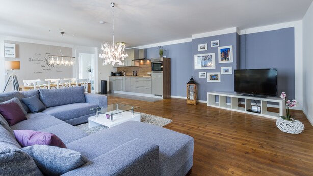 Una sala moderna con un sofá seccional gris, un televisor de pantalla plana, piso de madera y una cocina al fondo.
