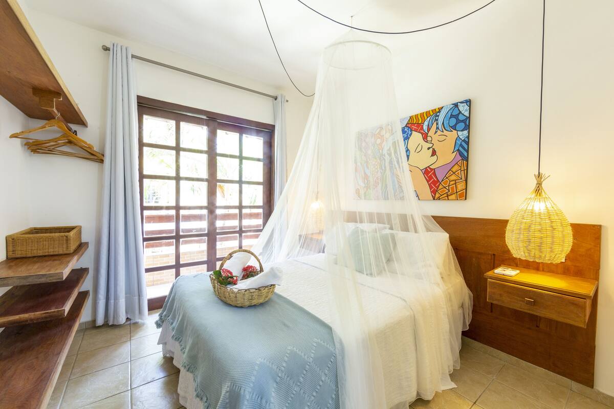 A bedroom with a bed, a bedside table, a hanging light, a window with curtains, a wall-mounted painting, and a wooden shelf with a basket.