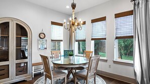 A dining room with a round glass table, wooden chairs, a chandelier, and large windows with blinds. - Bella Mar - Villas of Frangista, Community Pool, Walk to the Beach + Dining! (Miramar Beach)