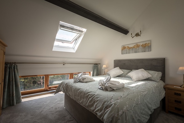 A bedroom with a large bed, a skylight, a view of greenery, and a nightstand with a lamp.
