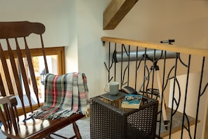 A wooden chair with a plaid blanket, a wicker chair, a small table with a mug, a book, and a telescope on a balcony railing.