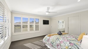 A bedroom with a bed, a ceiling fan, a TV, and large windows with blinds. - Guest House on Guy (Forster)