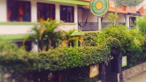 A building with a green and white facade, a sign with a yellow and green logo, and lush greenery around it.