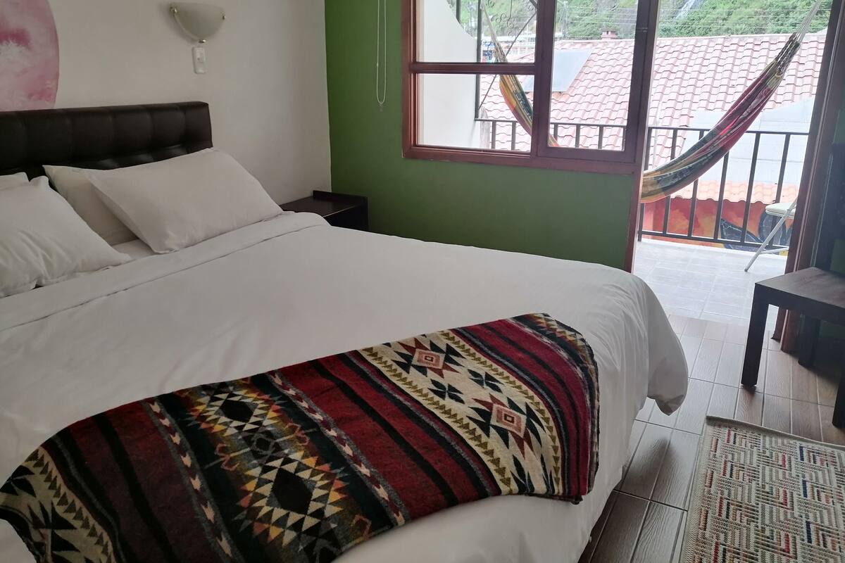 A neatly made bed with a patterned blanket, a bedside table, a wall-mounted lamp, and a view of a balcony with a hammock.