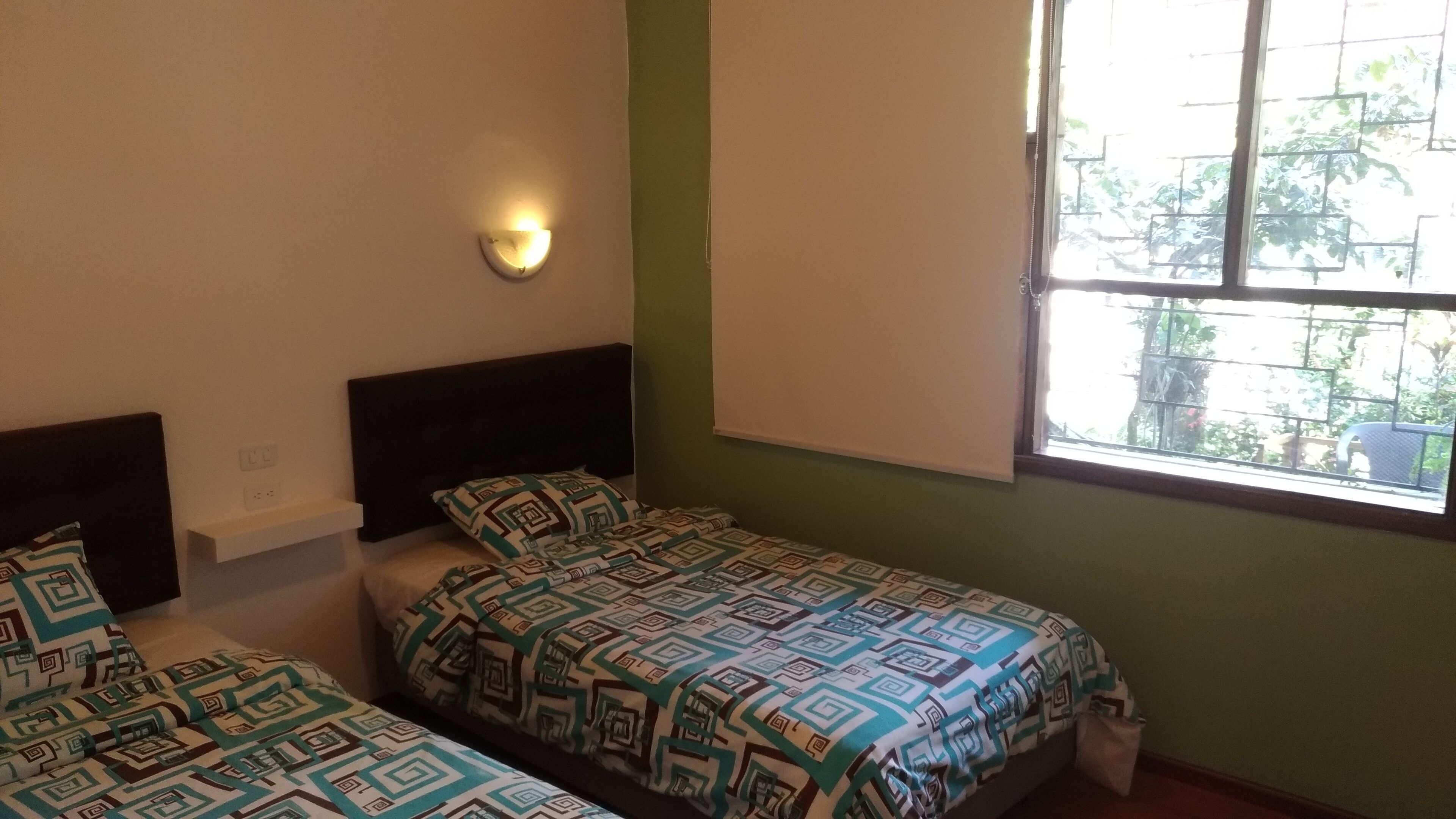 Una habitación con dos camas individuales, una lámpara fijada a la pared, una ventana con vistas a zonas verdes y una lámpara de techo blanca.