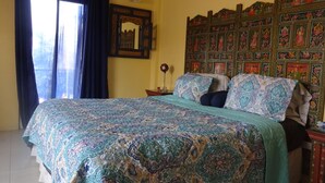 A bedroom with a large bed, a wooden headboard with artwork, a window with curtains, and a nightstand with a lamp. - Secret Haven TnT (Port of Spain)