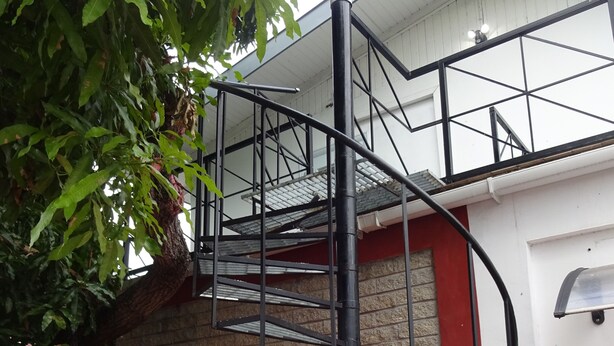 A building with a spiral staircase, a window with bars, and a tree in front. - Secret Haven TnT (Port of Spain)