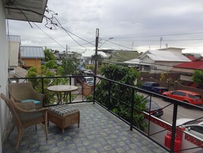 A balcony with wicker furniture, a tiled floor, and a view of a residential street with cars and houses. - Secret Haven TnT (Port of Spain)