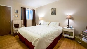 A bedroom with a large bed, a chair, a nightstand, a lamp, and a window with curtains.