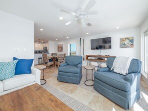 A modern living room with a white sofa, blue armchairs, and a coffee table. There is a dining area with wicker chairs and a TV mounted on the wall.