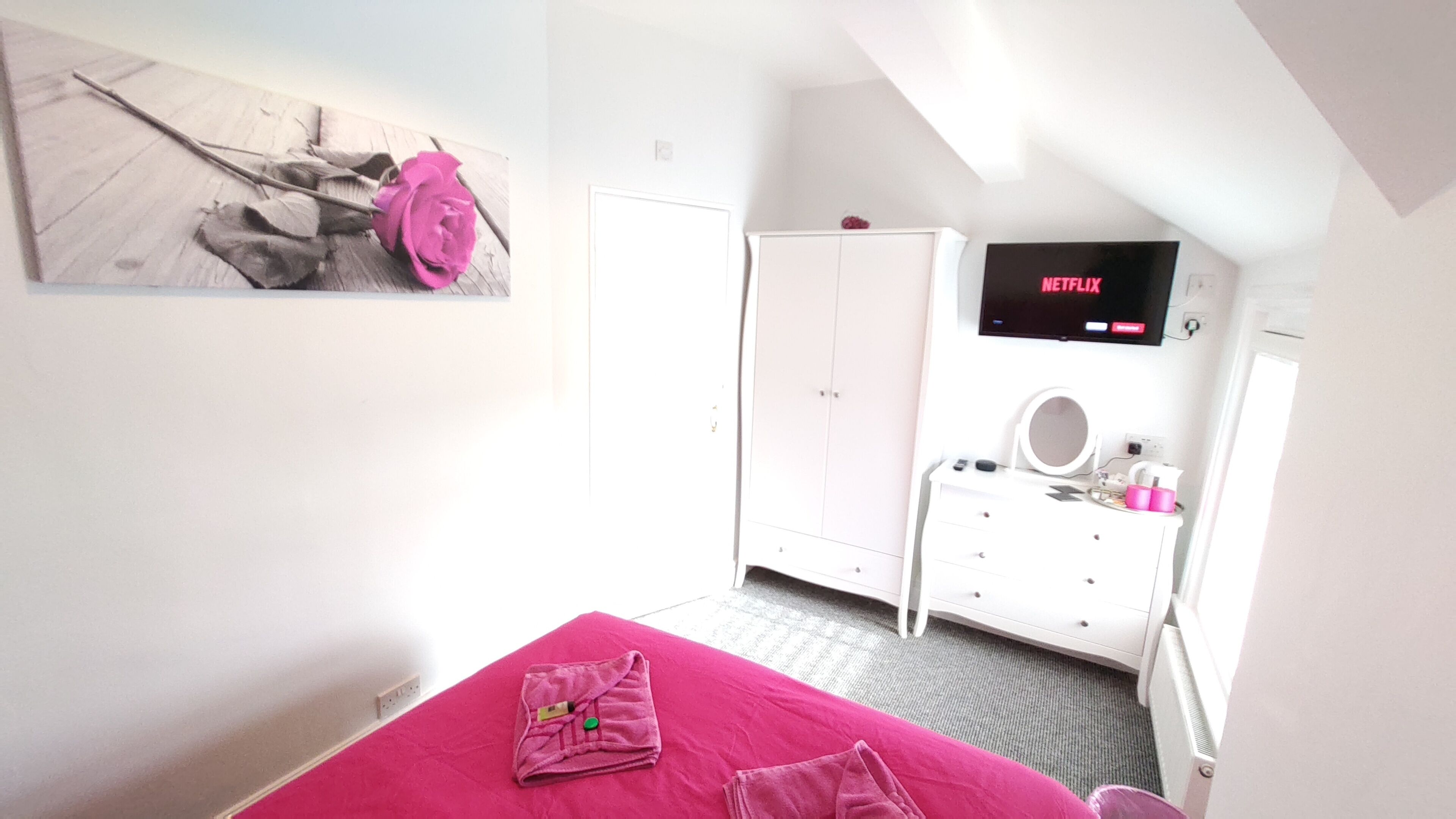 A bedroom with a bed, a wardrobe, a vanity with a mirror, and a television displaying 'NETFLIX'.