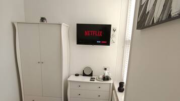 A hotel room with a bed, a wardrobe, a desk with a mirror, a coffee maker, and a wall-mounted TV displaying the Netflix logo.