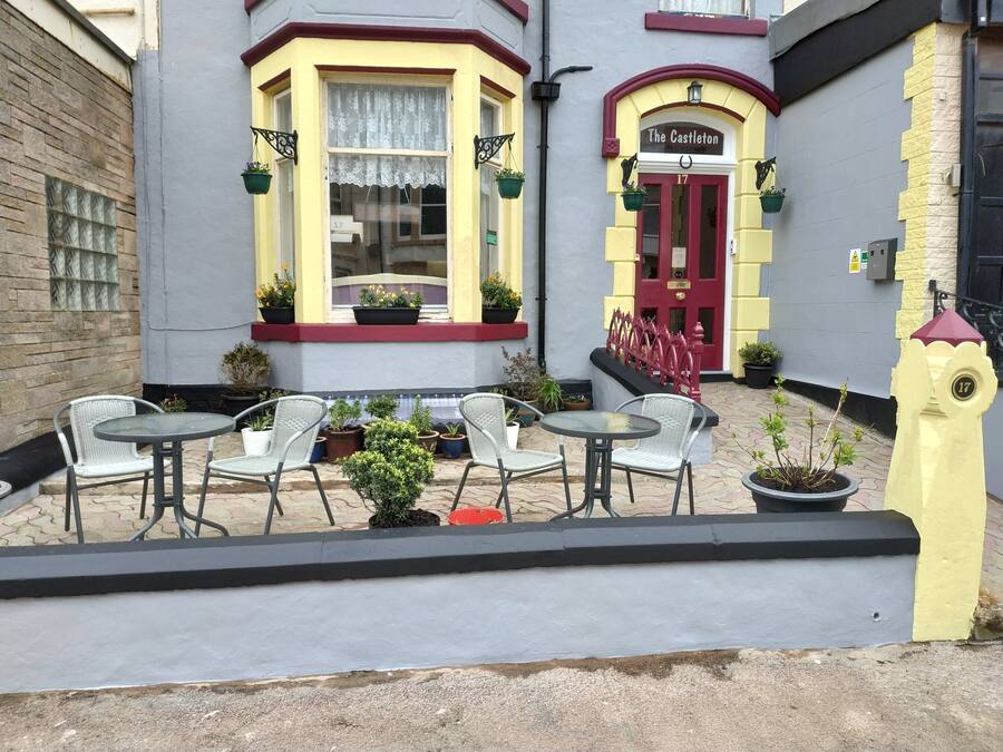A building with a red door labeled "The Castleton" and a number "17" on the wall. There are outdoor tables and chairs, and potted plants around the entrance.
