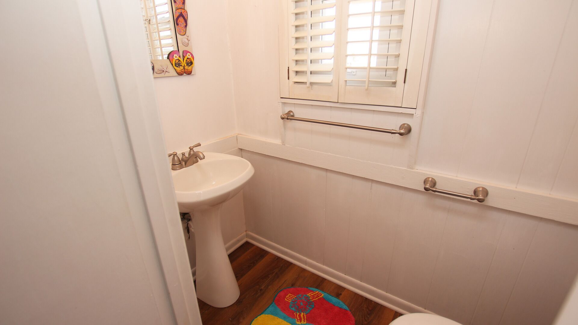 Une salle de bain avec un lavabo sur colonne, une fenêtre avec des stores, un porte-serviettes et un tapis coloré sur le sol.