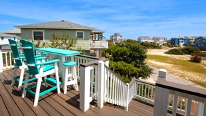 A wooden deck with turquoise chairs and a table, overlooking a beach with houses in the background. - Oceanside, Pool, Close to Beach, Sun Decks, OS1E, Seas The Day (Corolla)