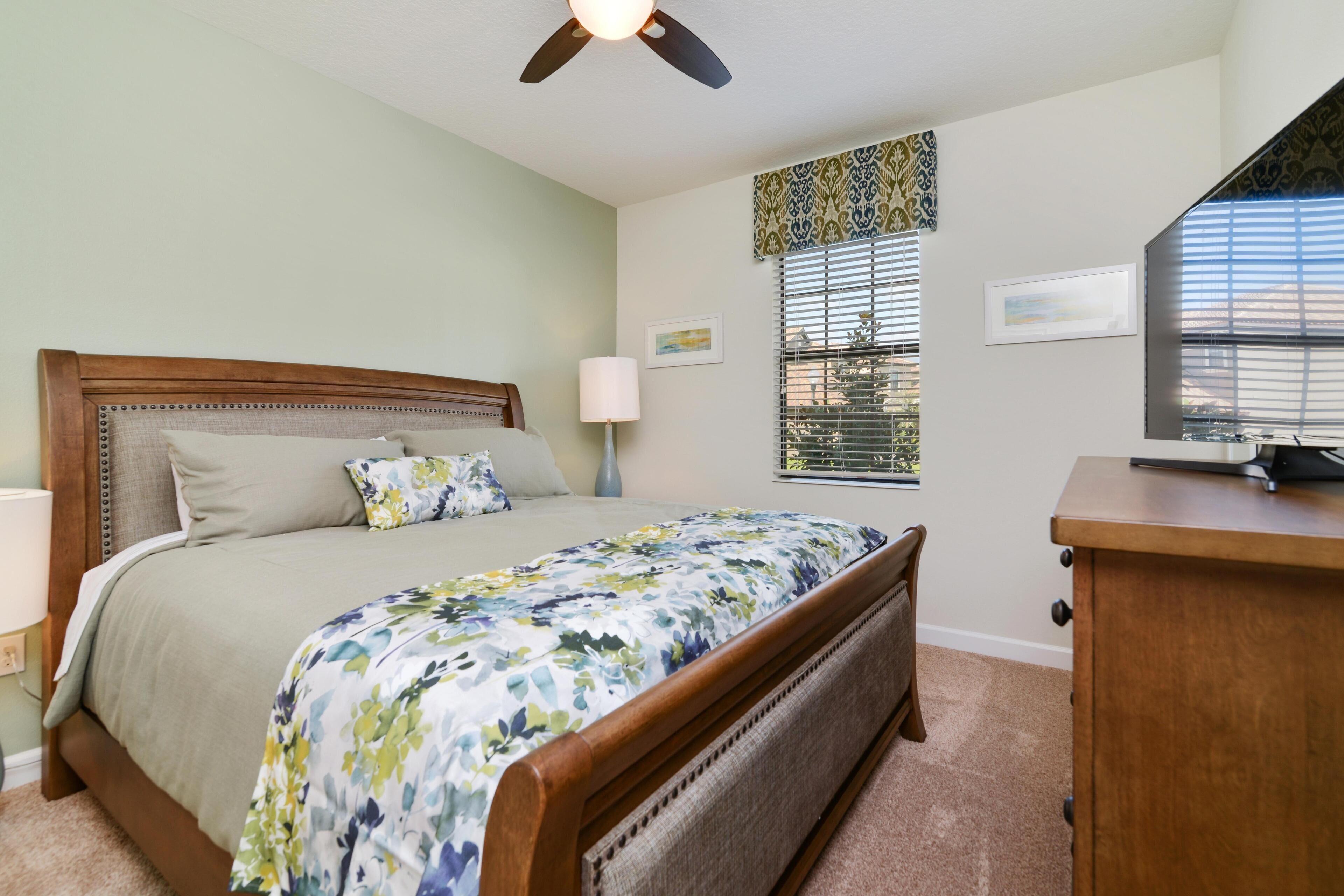 Une chambre à coucher comprenant un lit, une table de chevet, une commode, un ventilateur de plafond et une fenêtre avec des stores.