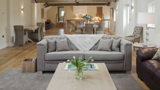 Ein geräumiges Wohnzimmer mit hoher Decke, freiliegenden Holzbalken, einer zentralen Leuchte, einem Sofa, einem Couchtisch und einem Essbereich im Hintergrund.
