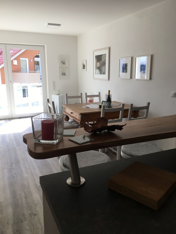 Un coin repas avec une table en bois, des chaises et un porte-bougie en verre. Des tableaux encadrés ornent le mur, et une fenêtre offre une vue sur les bâtiments à l’extérieur.