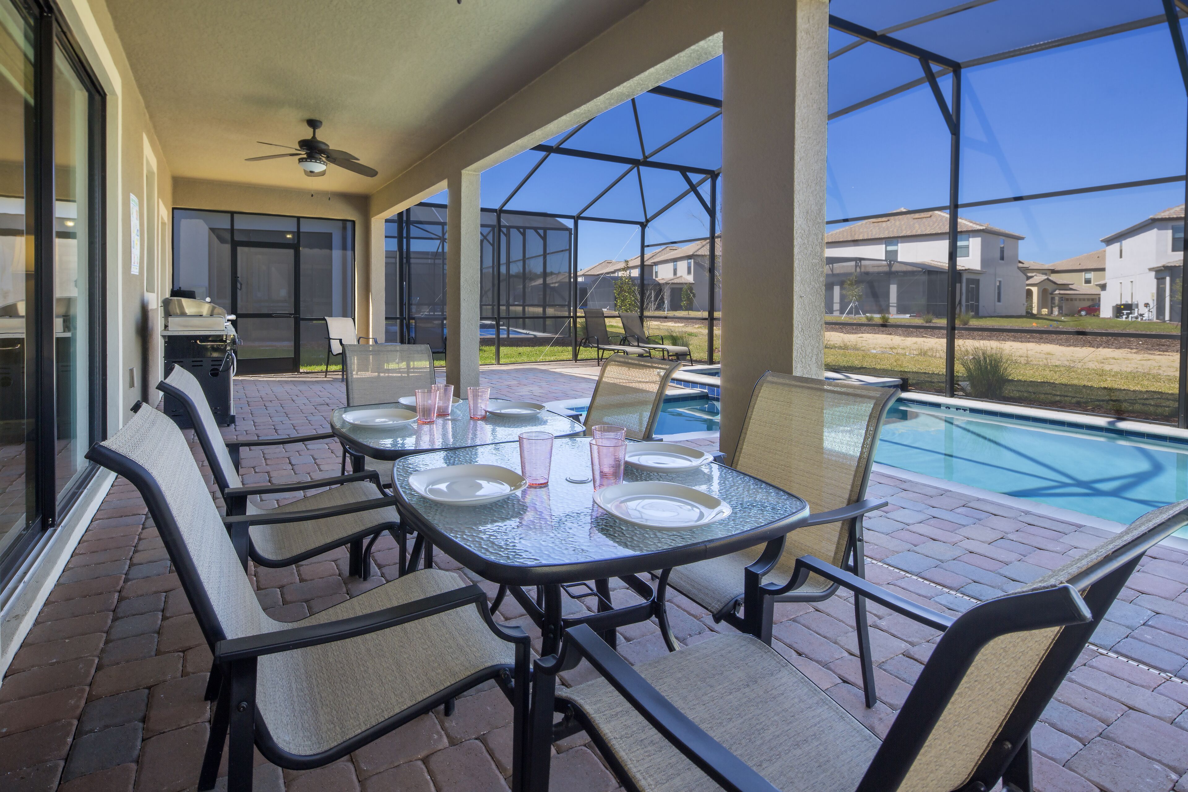 Un patio techado con una mesa lista para comer, sillas y vistas a una piscina y casas al fondo.