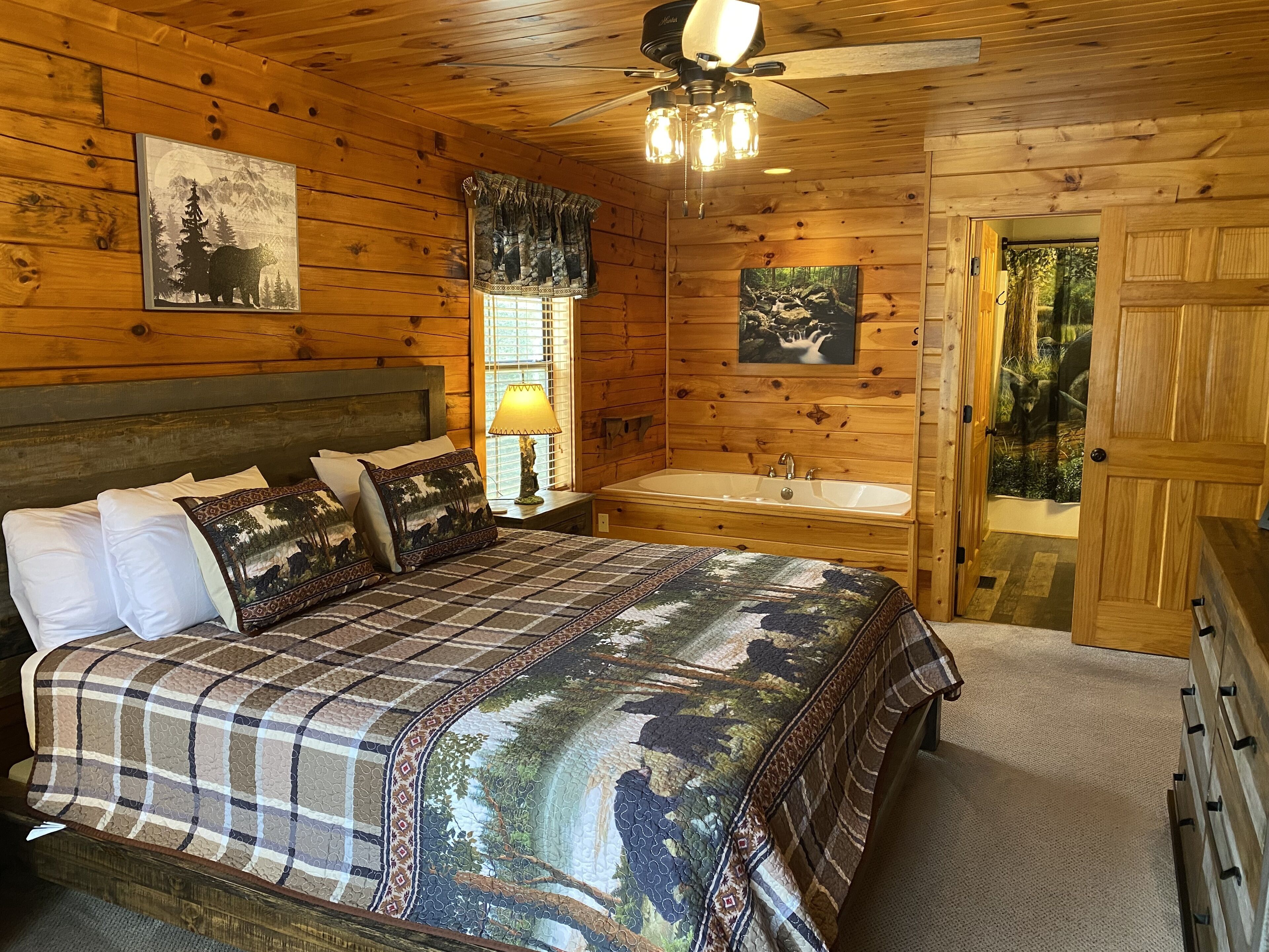 A wooden cabin bedroom with a bed, a nightstand, a window with curtains, a wall-mounted painting, a ceiling fan, and a door leading outside.