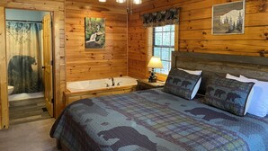 Ein Schlafzimmer in einer Holzhütte mit Bett, einem Fenster mit Blick auf Bäume, einem Deckenventilator und einem Badezimmer mit Badewanne.