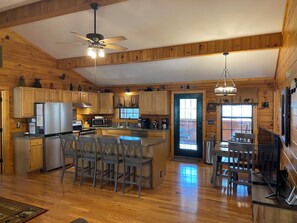 Une cuisine spacieuse avec des meubles en bois, un îlot central doté de tabourets de bar, un ventilateur de plafond et une vue sur l’extérieur grâce à une porte vitrée.
