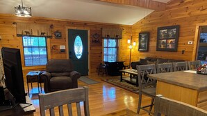 Ein gemütliches Blockhauss-Interieur mit Kamin, Holzwänden, einer Essecke und einem Wohnbereich mit Sofa und Sesseln.