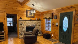 Ein gemütliches Blockhaus-Interieur mit Kamin aus Stein, Ledersitzmöbeln, einem Fernseher und einer Tür mit einem dekorativen Glasfenster.