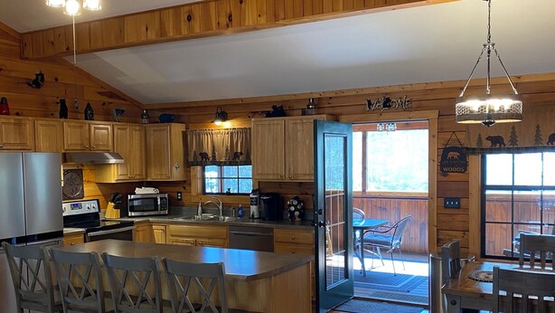 Eine geräumige Küche mit Holzausstattung, einer zentralen Kochinsel inklusive Barhocker, einem Deckenventilator und Blick ins Freie durch eine Glastür.