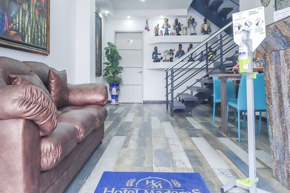 A hotel lobby with a leather sofa, a blue rug with 'Hotel Madras' logo, a staircase, and various decorative items on shelves. - Hotel Los Maderos (Cali)